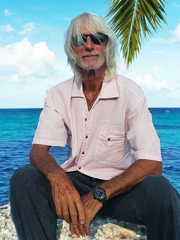 Camicia maniche corte Un uomo con barba e capelli lunghi bianchi è seduto su un grosso sasso, alle spalle ha un mare caraibico con una foglia di palma nel cielo azzurro, indossa una bella camicia maniche corte