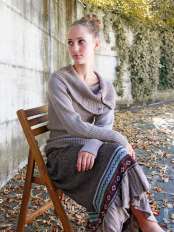 Donna bionda seduta su una sedia, con maglione e gonna ampia in lana di alpaca, gambe accavallate, modello New Folk.