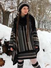 Donna con trecce in paesaggio innevato, indossa il poncho di alpaca a motivo jacquard, base nera, con basco, gonna e scaldamuscoli coordinati.