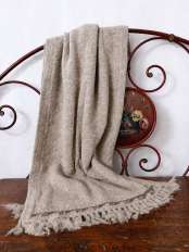 Scialle da donna beige in lana di alpaca con frange, appoggiato a una spalliera di letto in ferro su legno antico, modello Kora.