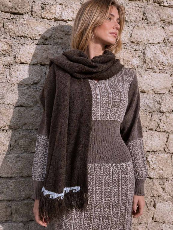 Donna bionda davanti a un muro in pietra con vestito corto e uno scialle da donna in lana di alpaca marrone con frange, modello Kora.